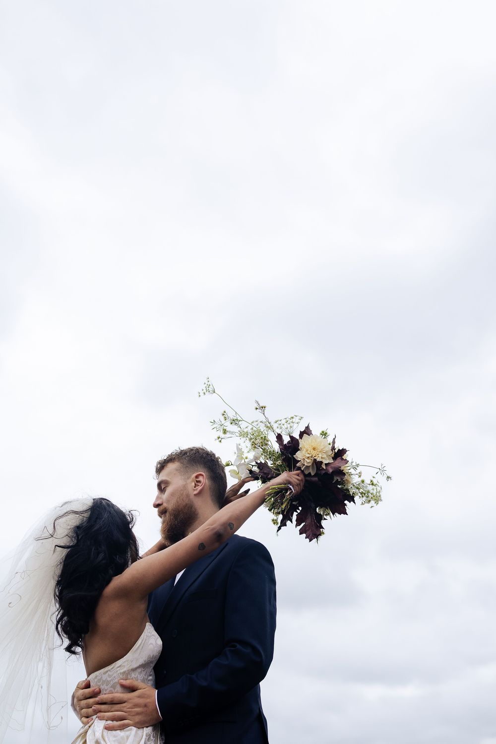 Romantic wedding couple embraces outdoors against cloudy sky with bride holding floral bouquet.