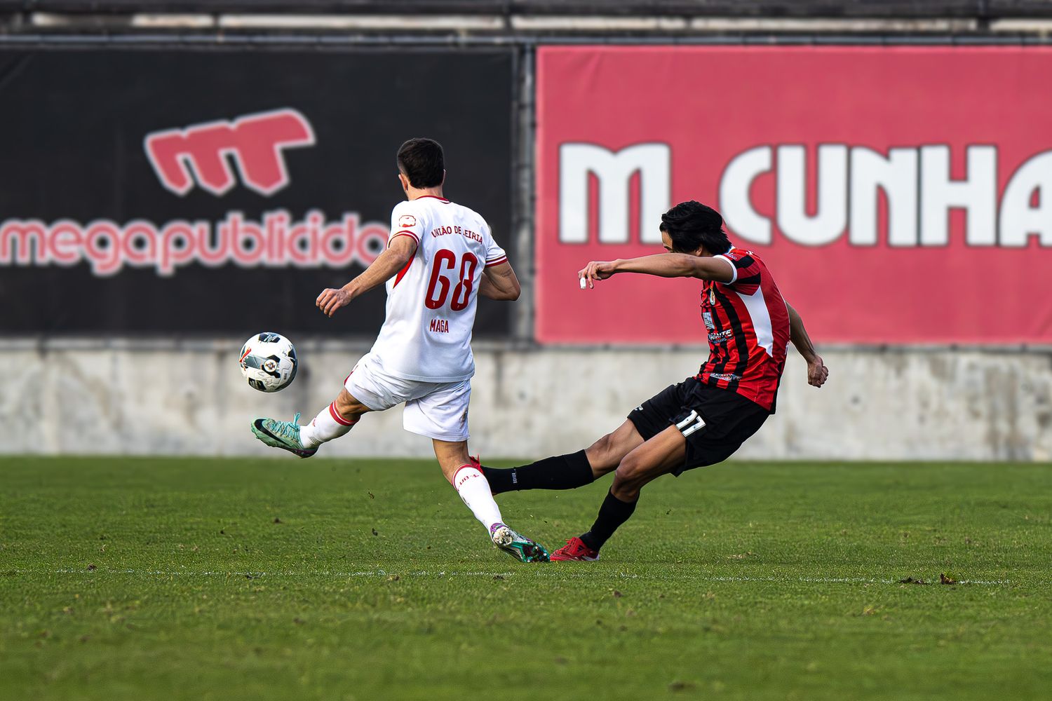 Soccer action shot with players sliding on grass field near advertising boards.
