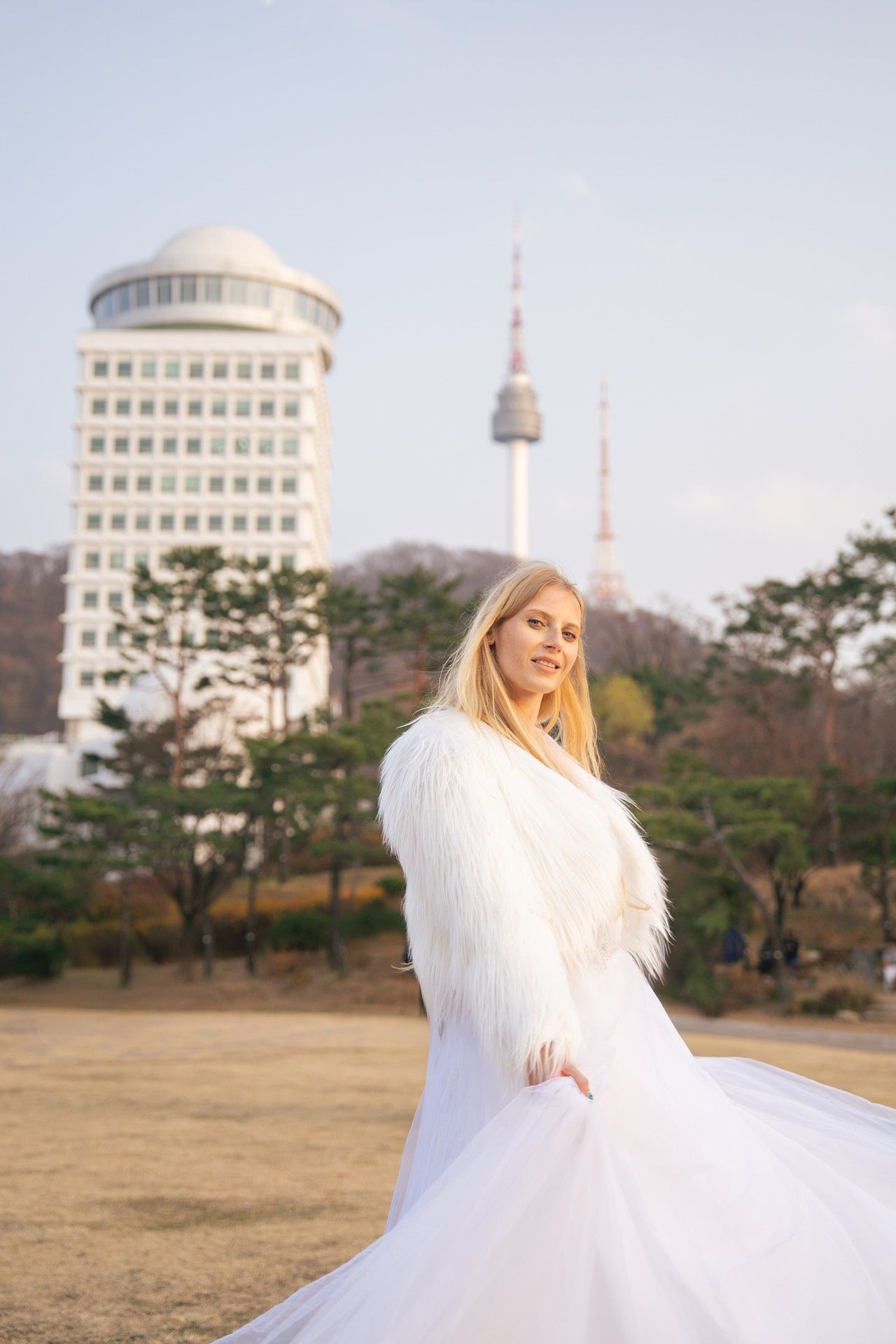 A figure in a flowing white dress moves gracefully across an open field with city buildings and a tower in the background.