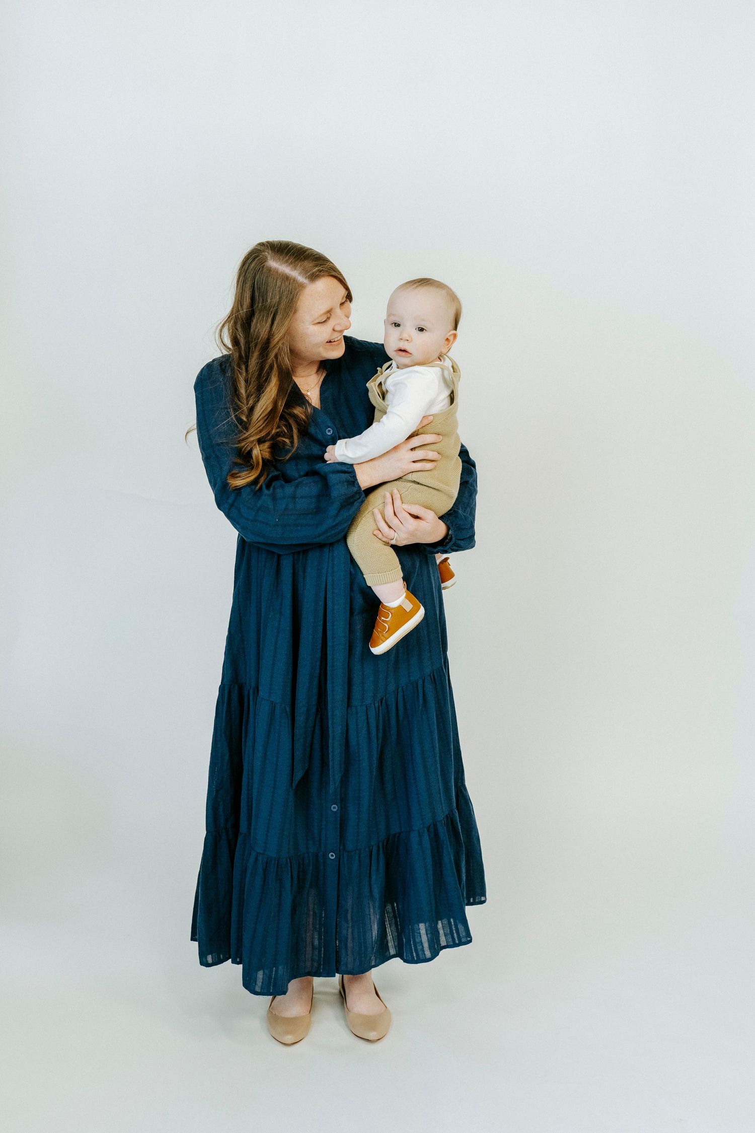A parent in a navy tiered maxi dress holds a small baby wearing white against a light background.