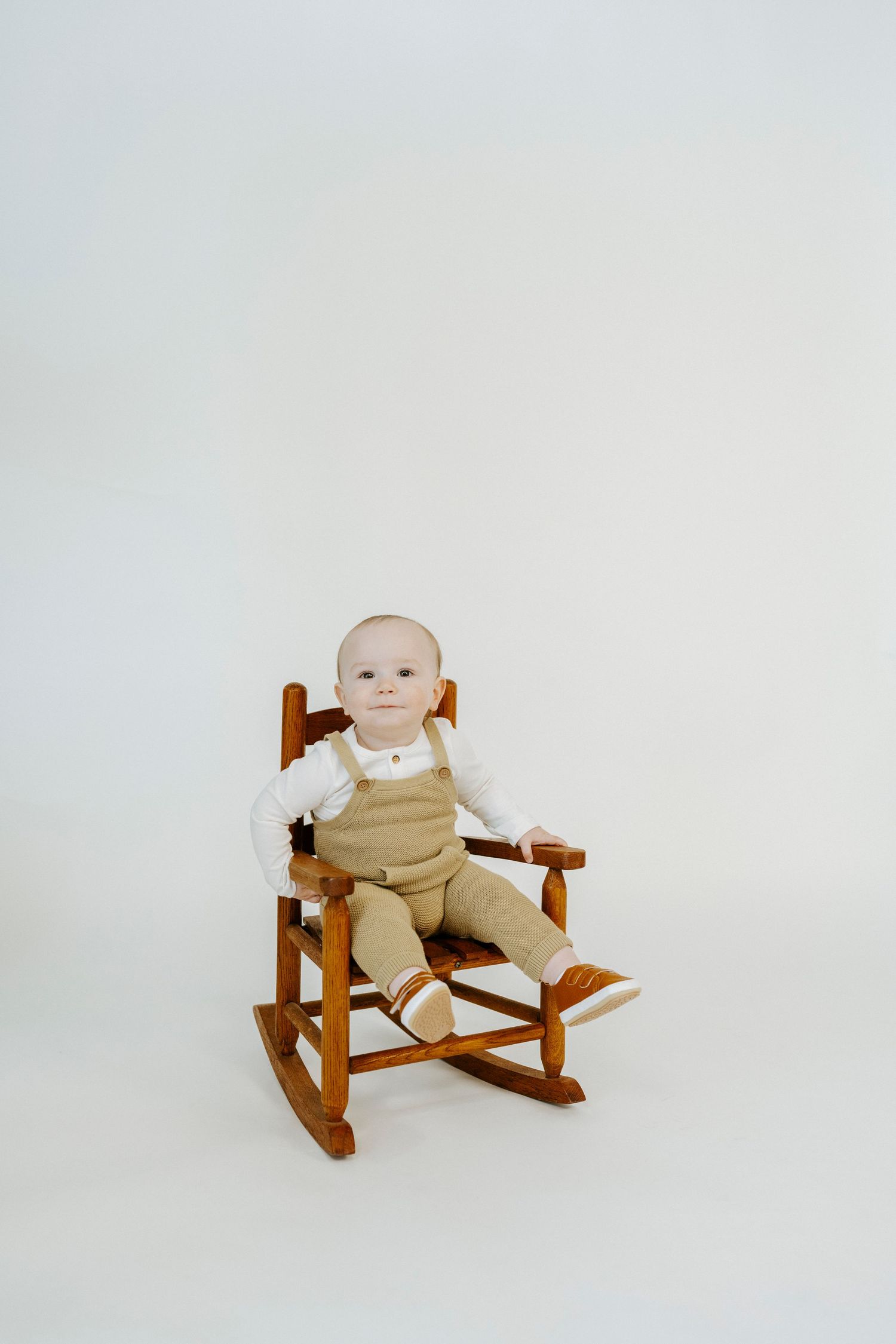 A baby wearing beige overalls sits in a wooden rocking chair against a plain white background.