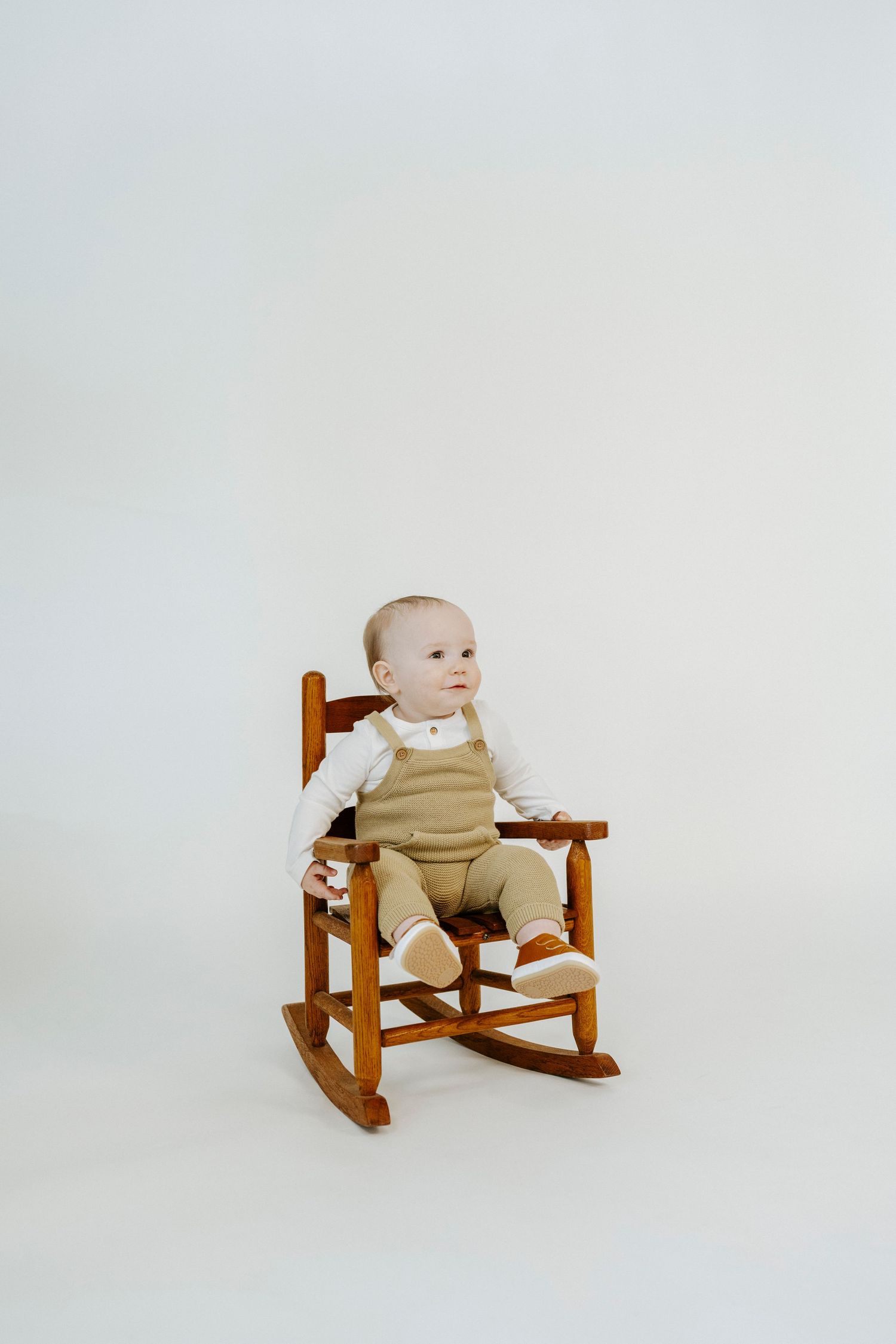 A small baby sits in a wooden rocking chair wearing beige clothing against a white background.
