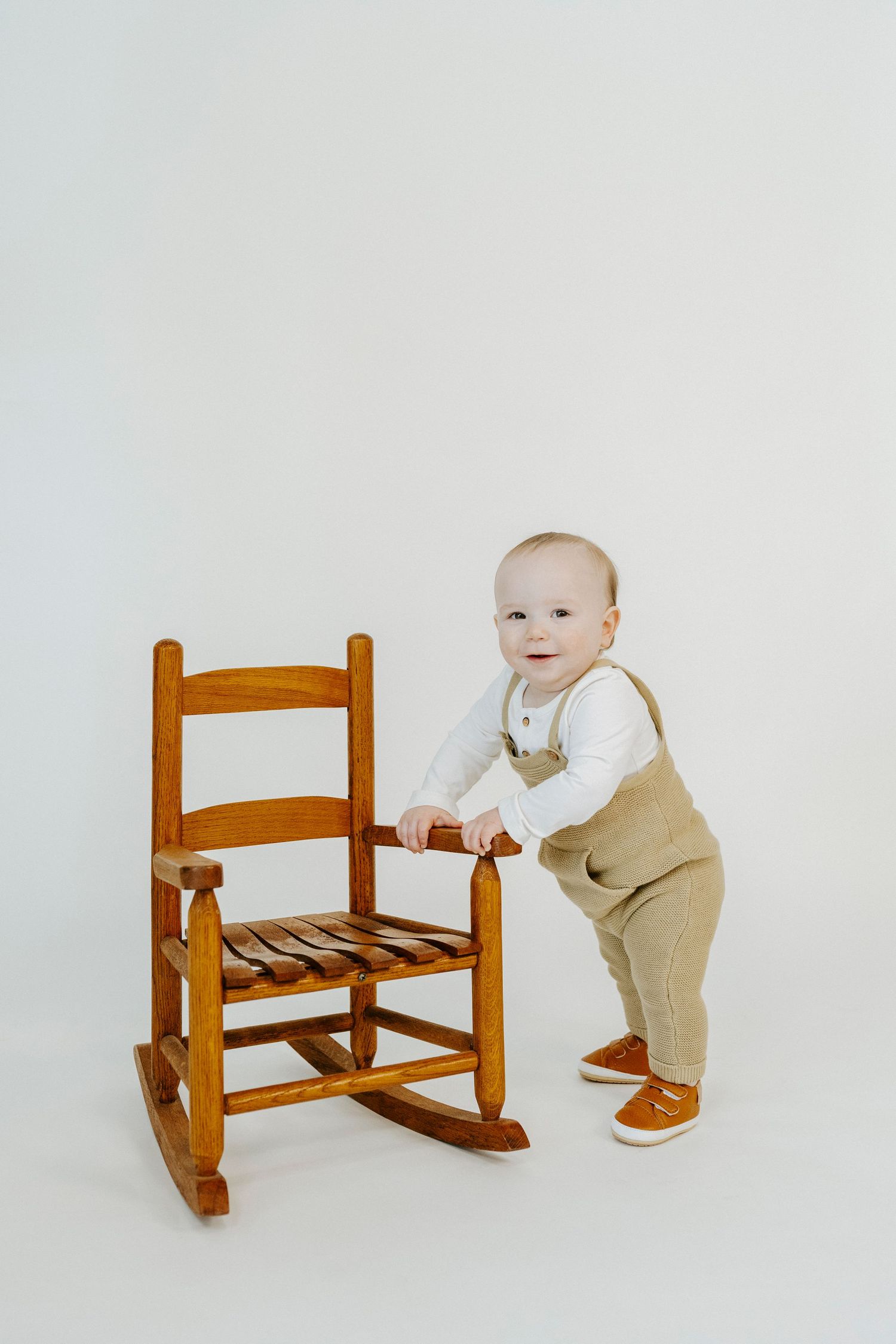 Small child in khaki overalls stands beside wooden rocking chair in minimalist studio setting.