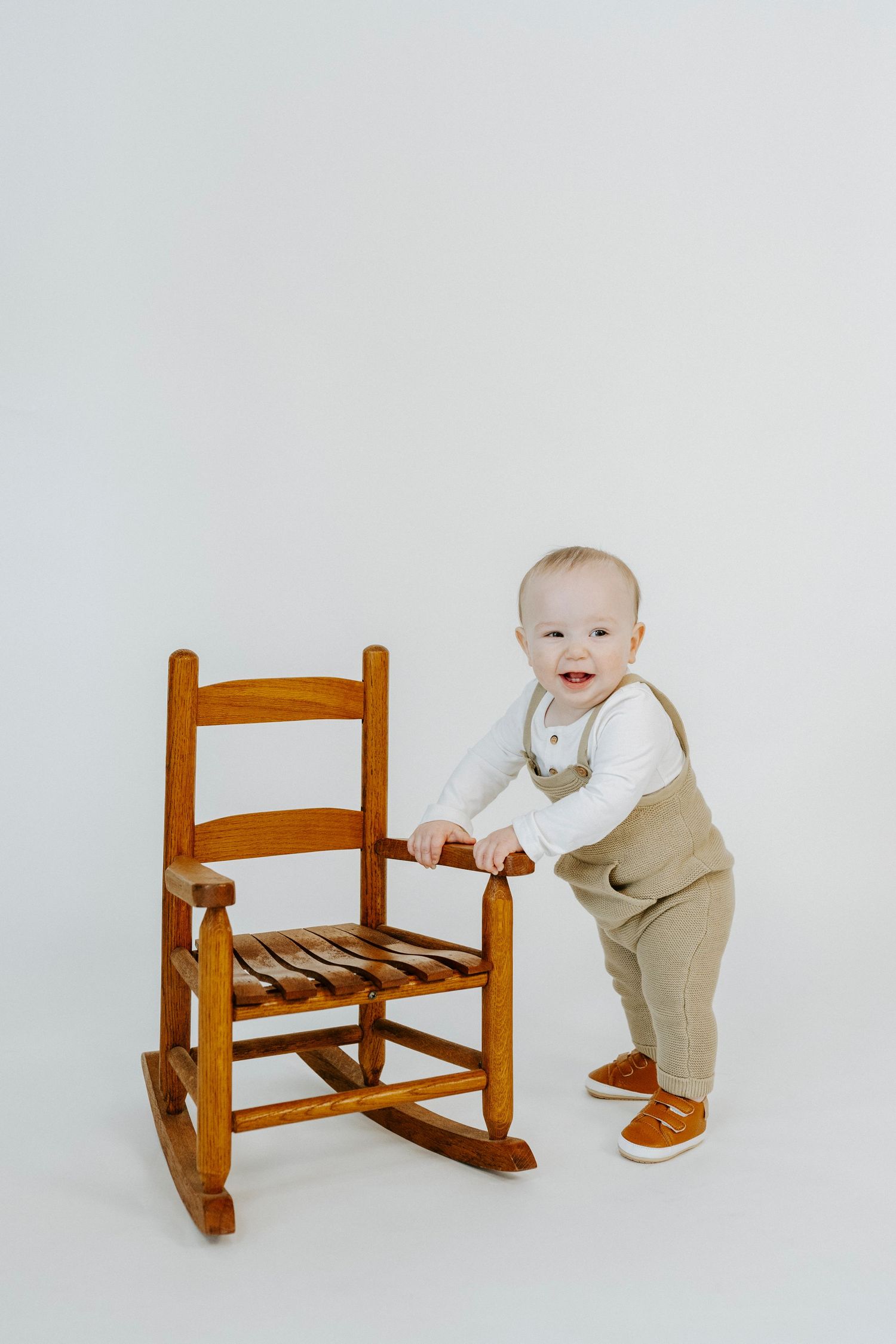 Baby in beige overalls and white shirt smiles while holding onto wooden rocking chair.