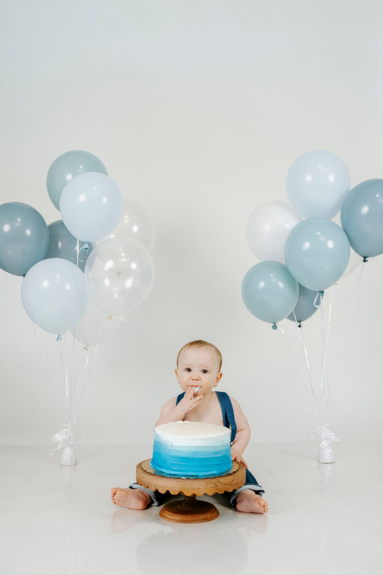 Baby's first birthday cake smash photo session with light blue and white balloons against white backdrop.