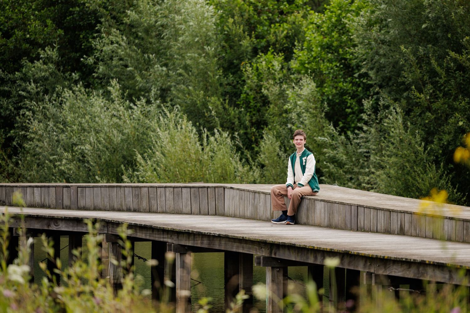 A person in a blue shirt walks across a wooden boardwalk path surrounded by lush green foliage on a sunny day.