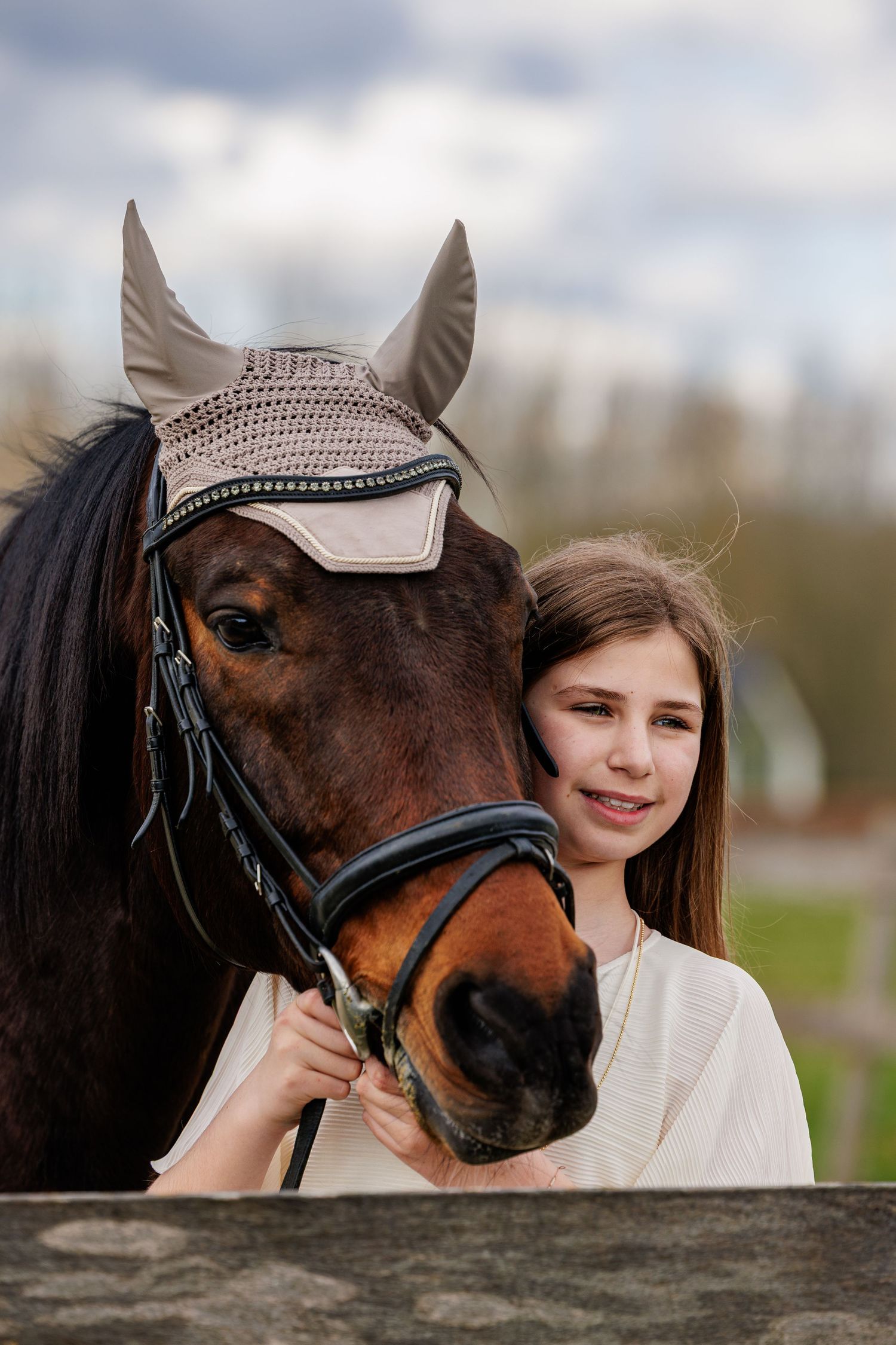 A brown horse wearing a decorative bridle and bonnet poses with someone in a white top against a cloudy sky.