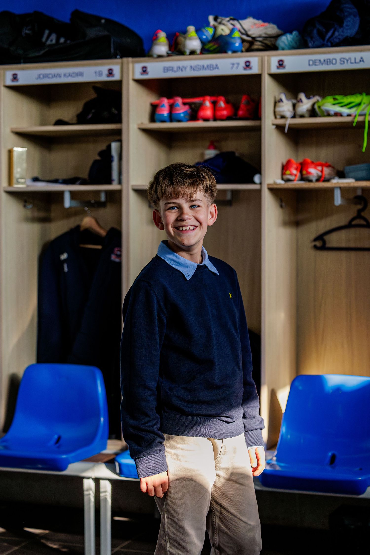 Child sits on a blue bench in front of storage lockers in what appears to be a sports locker room or changing area.