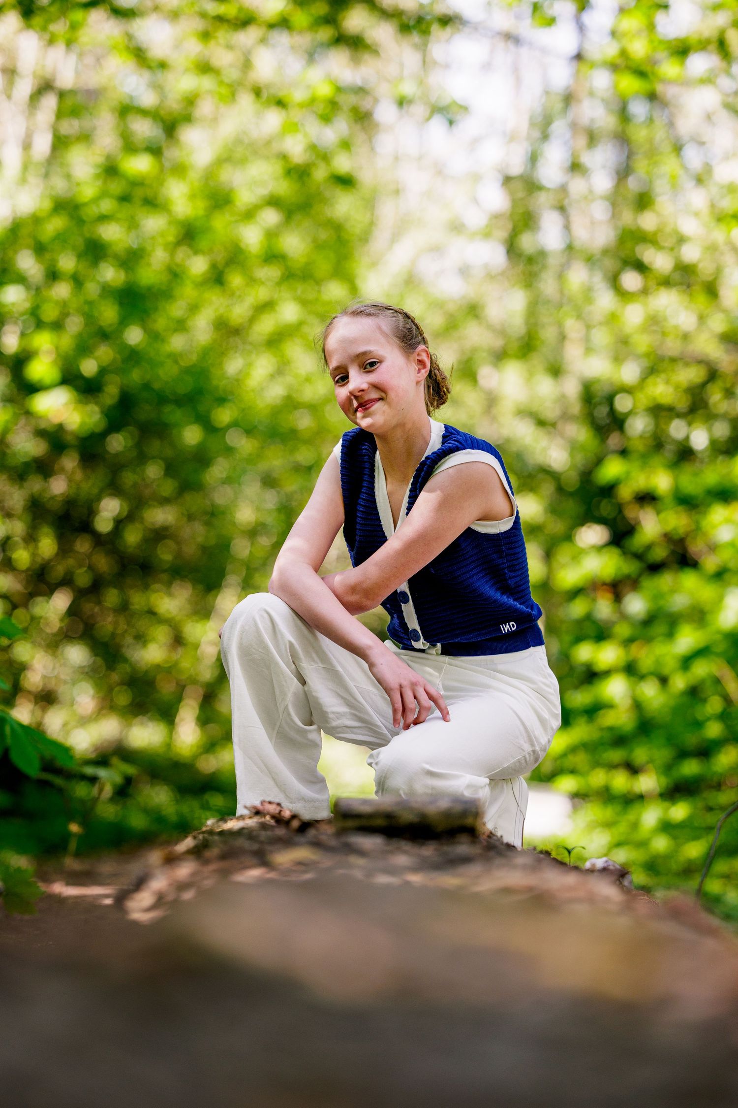 Person in navy sleeveless top and white pants sits casually on a rock in a sunlit forest setting.