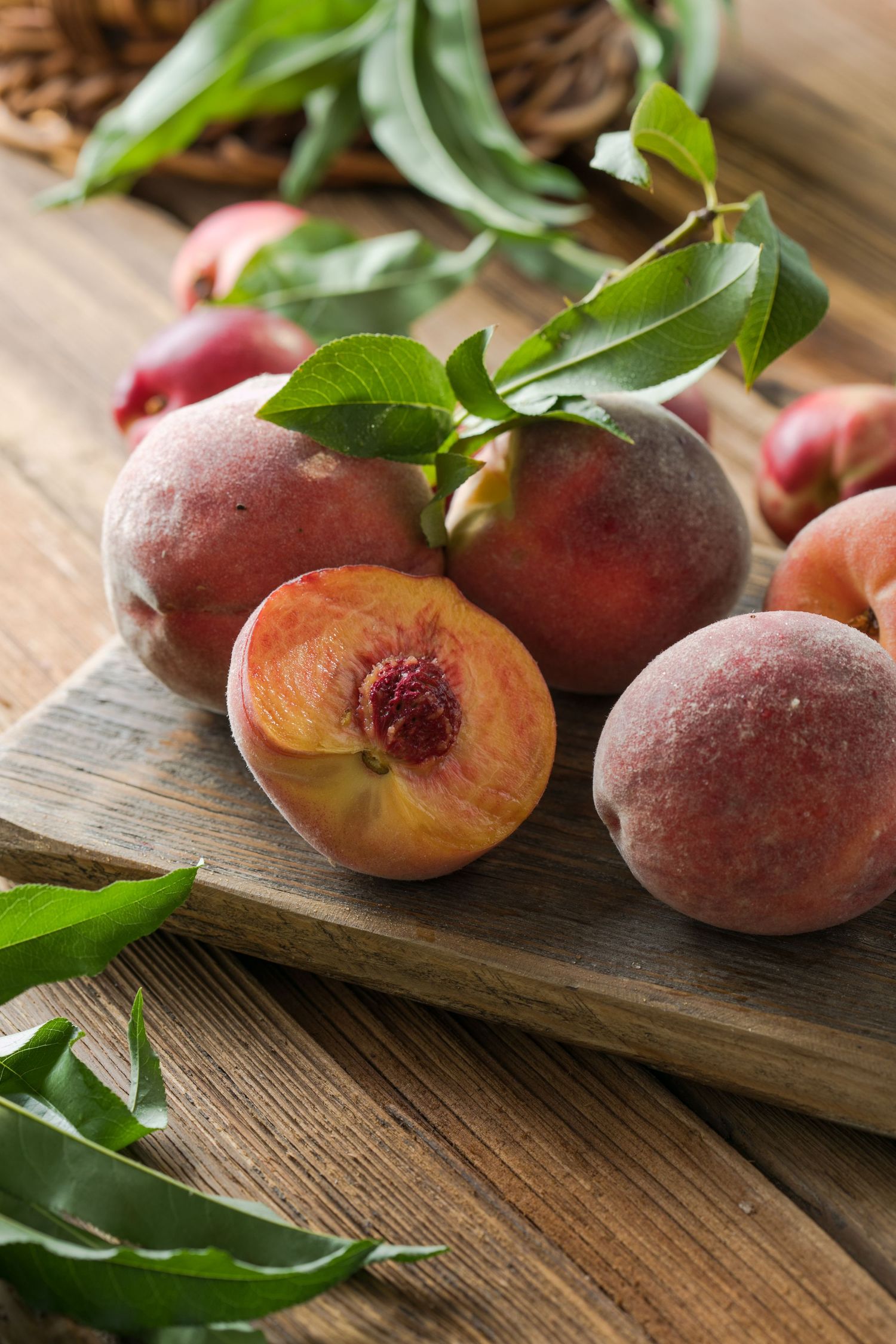 Fresh ripe peaches with green leaves arranged on a rustic wooden cutting board showing whole and halved fruit.