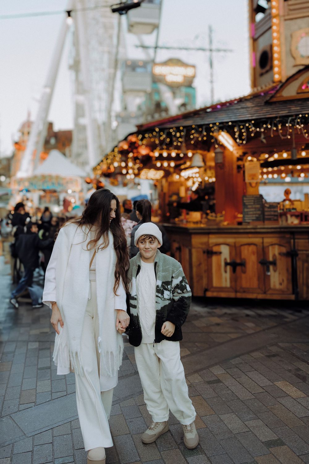 People in matching white outfits walking through a festively lit winter market with wooden stalls.