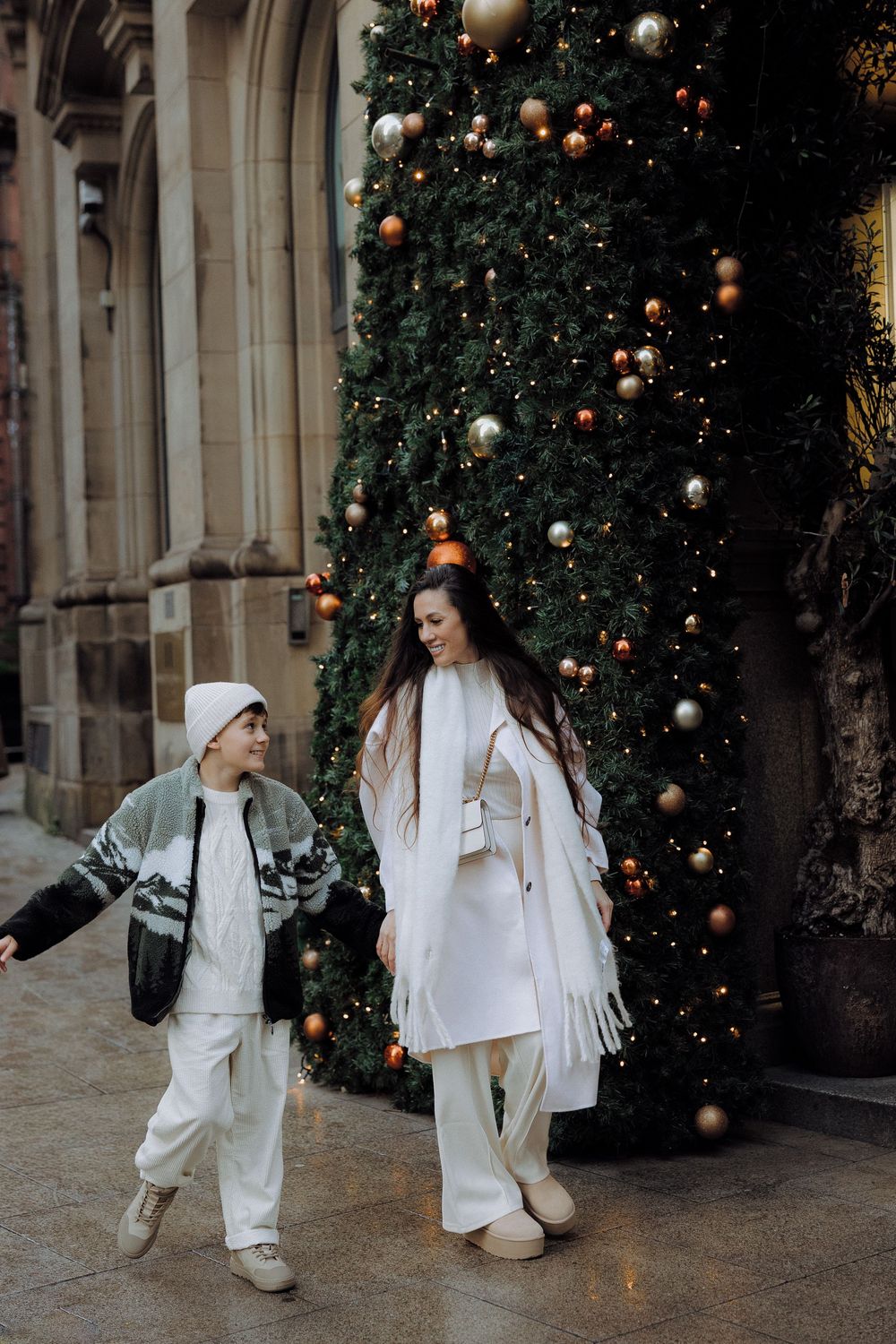 Two figures in white outfits pose playfully near a decorated Christmas tree with ornaments and lights.