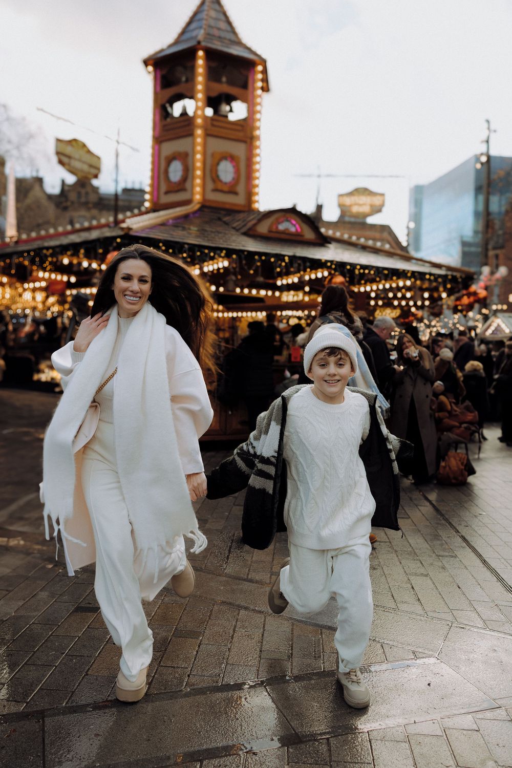 People in white winter outfits run joyfully through a festive Christmas market with twinkling lights.