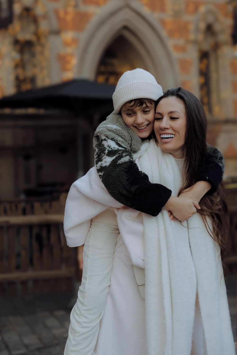 Two people in cozy white winter outfits sharing a joyful moment against a brick building backdrop.