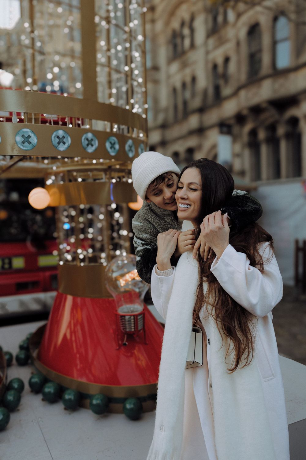 Two people share a joyful moment near a festive carousel with twinkling lights and holiday decorations in an urban setting.