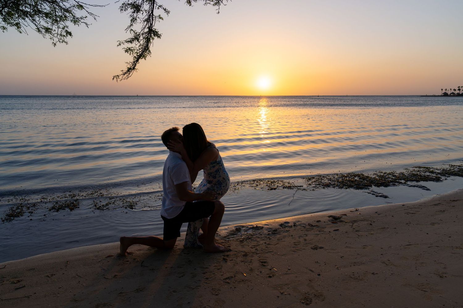 Silhouettes of people watching a vibrant orange sunset over calm ocean waters from a sandy beach.