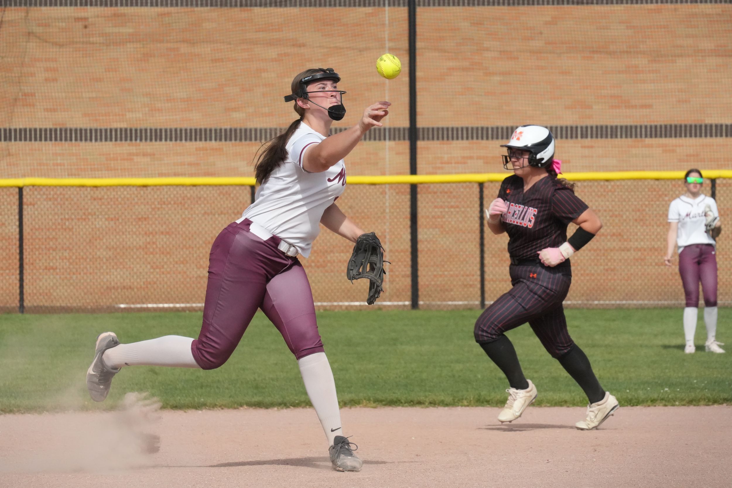 Softball player in maroon uniform throwing ball during game while teammate runs bases on sunny day.