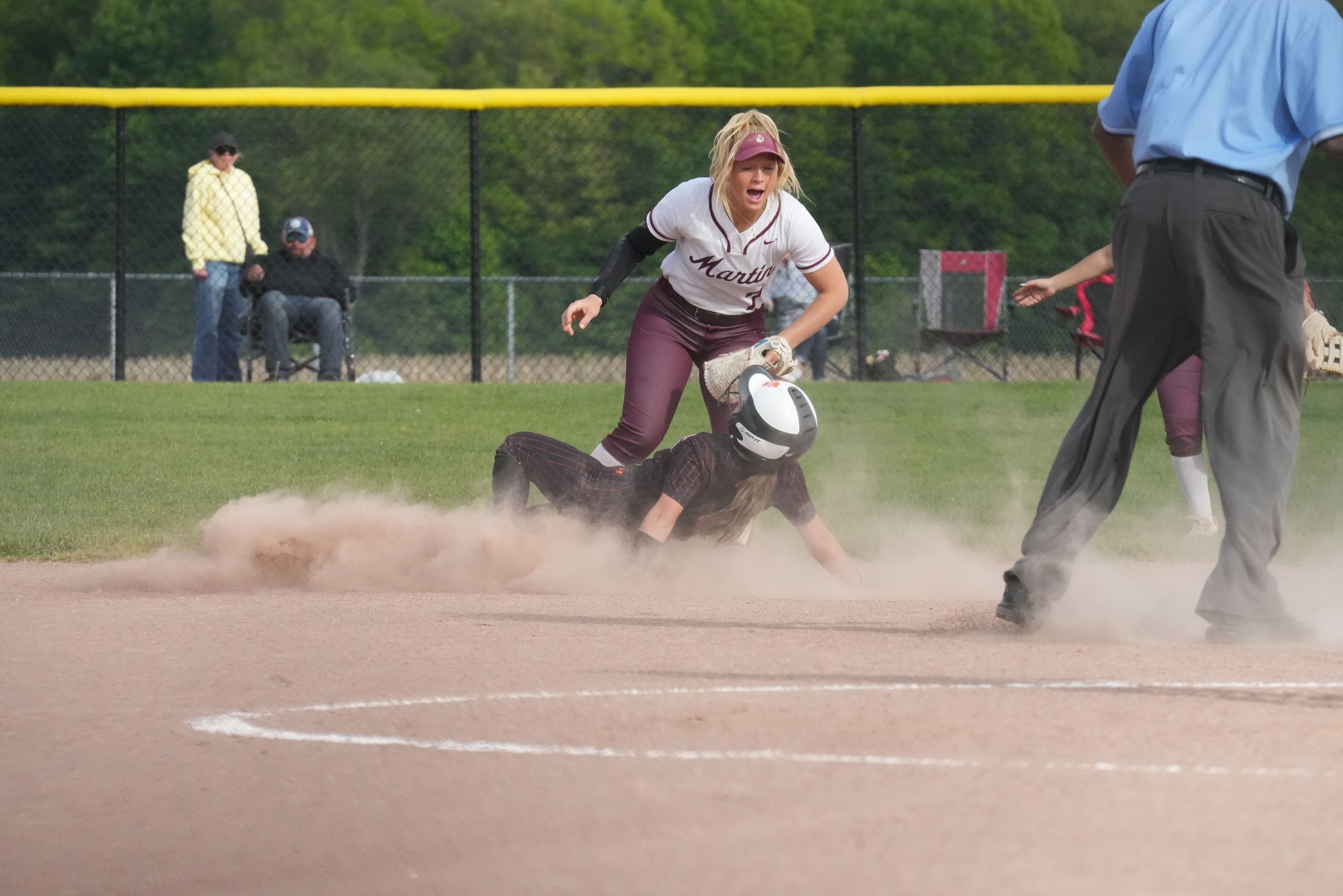 Softball player tags base in cloud of dust during intense game play, with umpire watching closely.