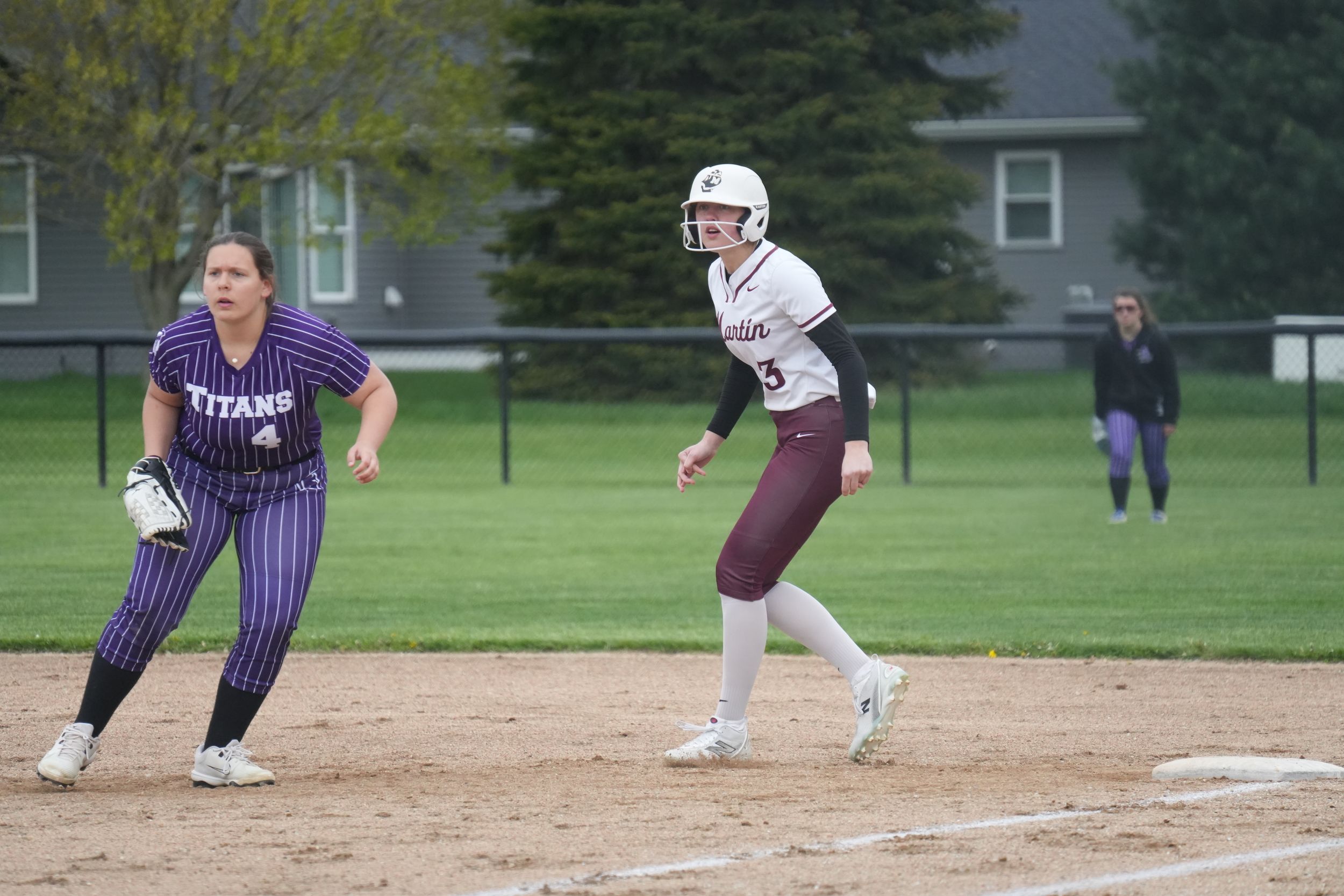 Softball player in maroon uniform rounds base as purple Titans fielder watches during a game on a dirt infield.