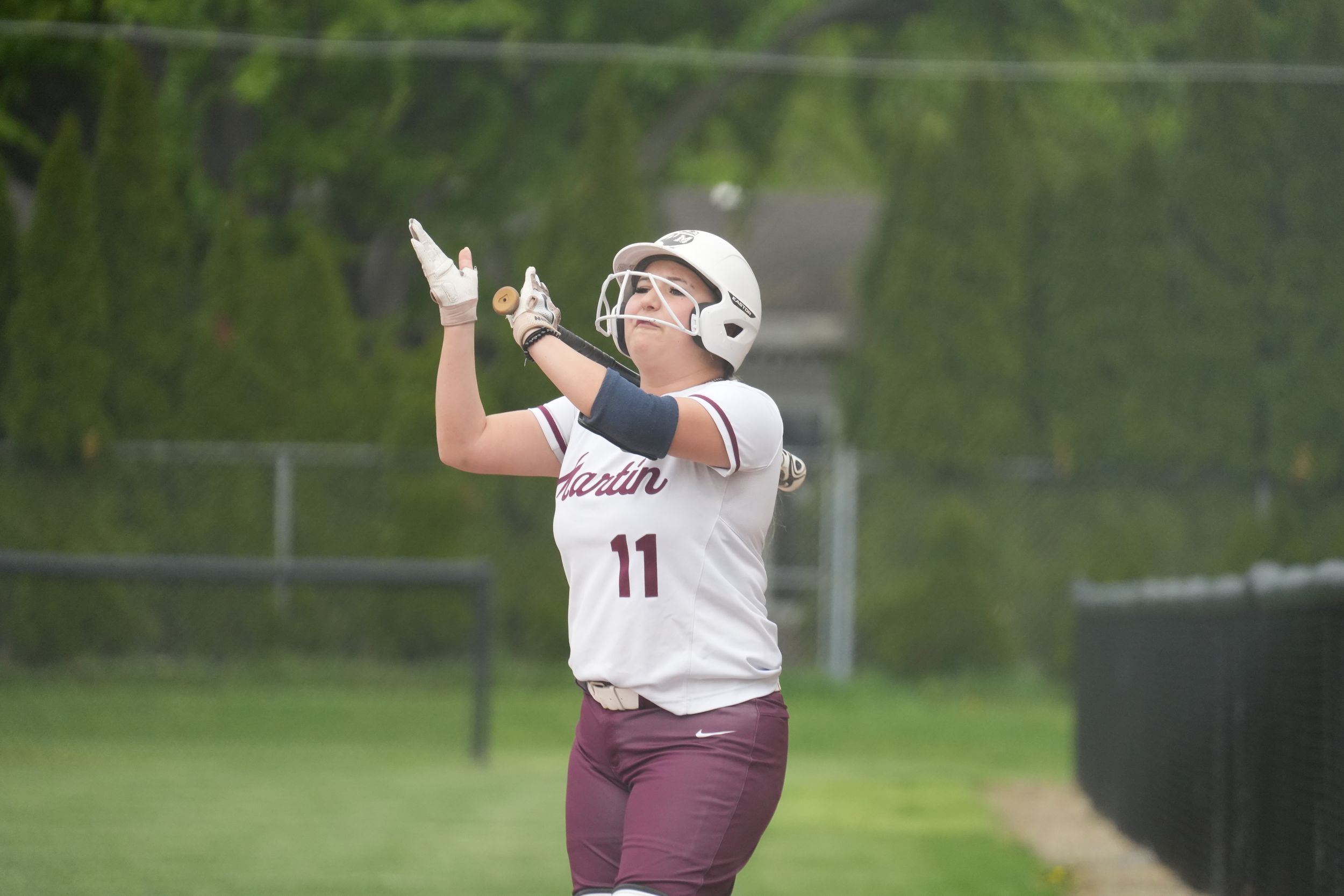 Softball player #11 in maroon uniform celebrates with hands raised after hitting, wearing helmet on outdoor field.