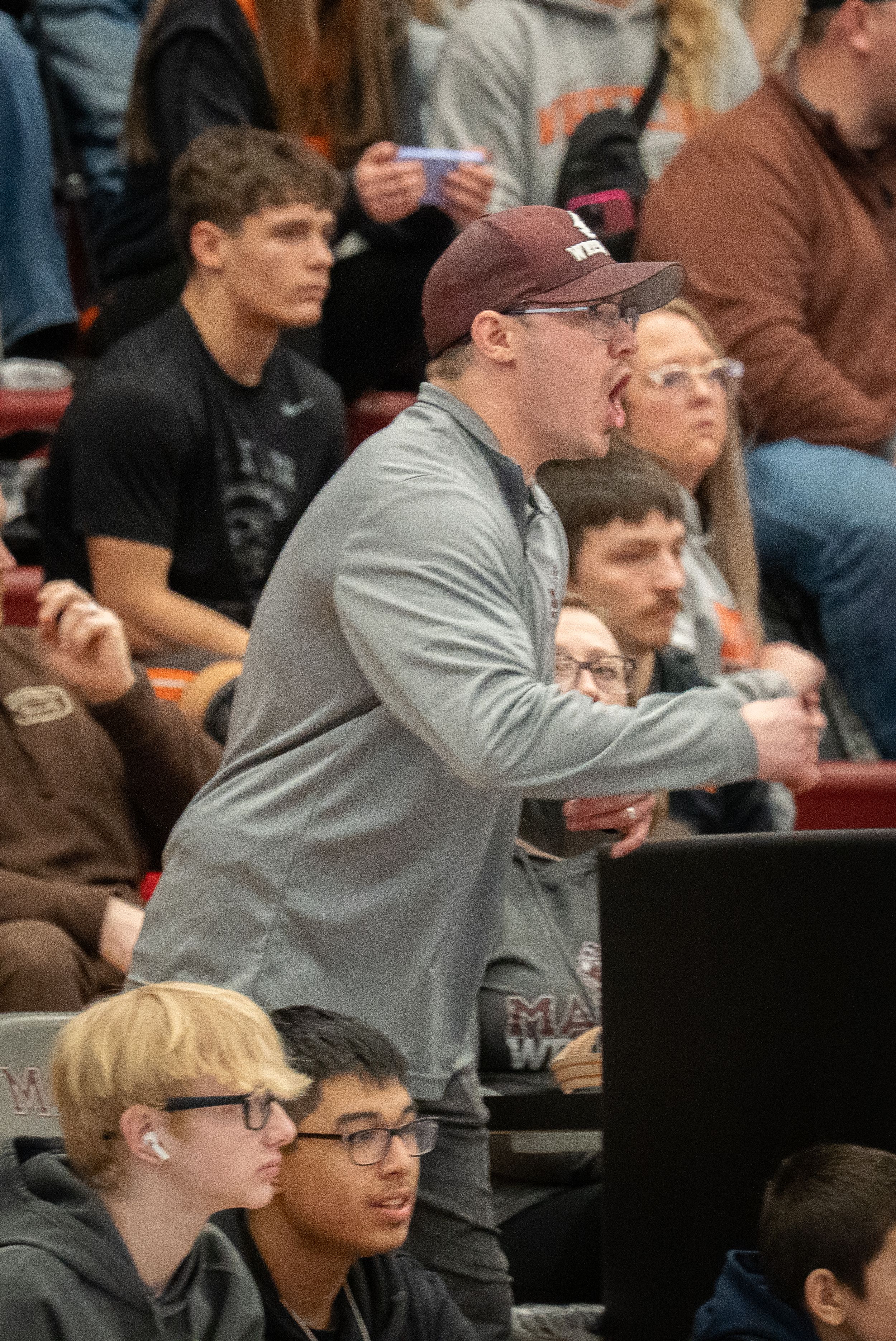 A man in a gray jacket and maroon cap shouts instructions courtside at an indoor basketball game with fans in the stands.