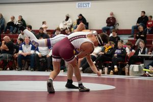 Two wrestlers compete on a mat during an indoor wrestling match with spectators watching in the background.