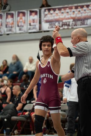 A wrestler in a maroon Martin singlet has his arm raised by a referee after winning a match at a wrestling tournament.