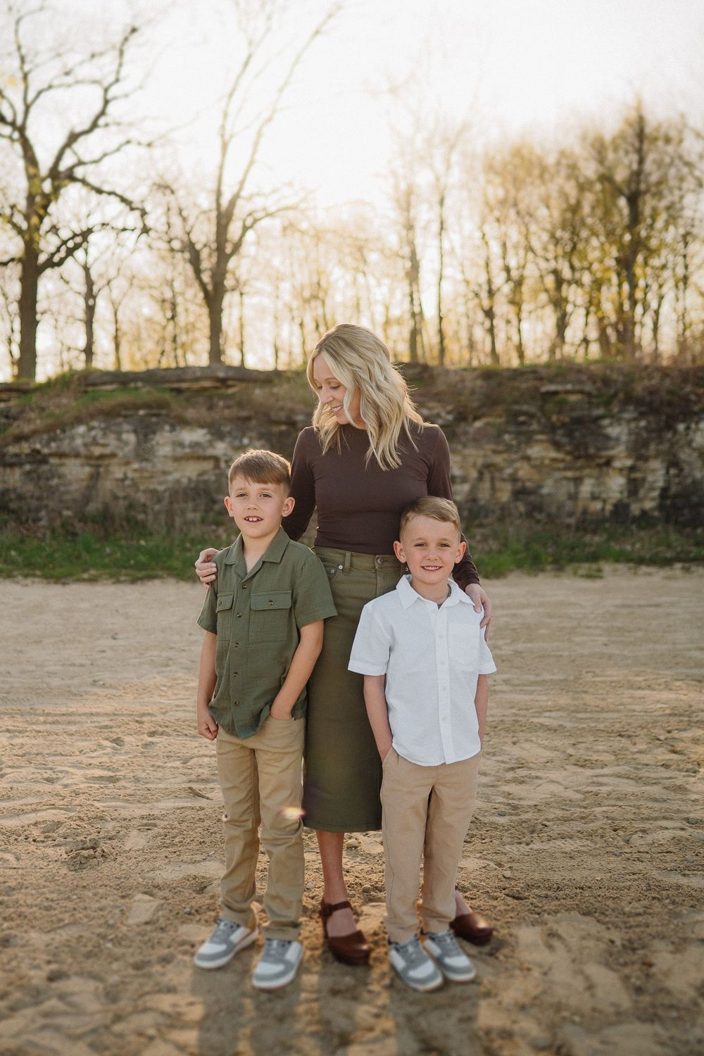 Family group wearing coordinated earth-tone outfits poses together on dirt path with stone wall and autumn trees behind them.