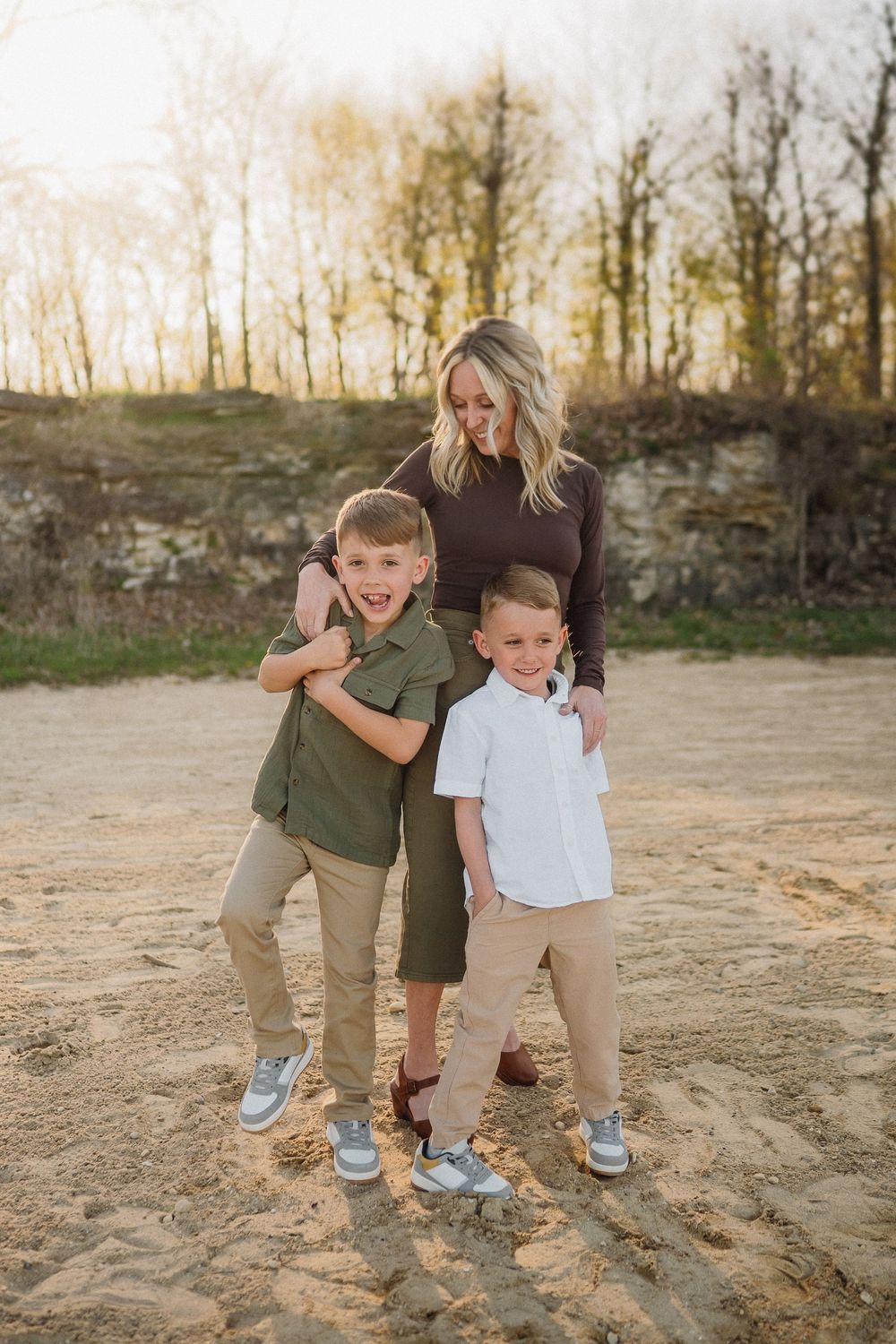A family of three poses together outdoors at sunset in front of a stone wall and trees.