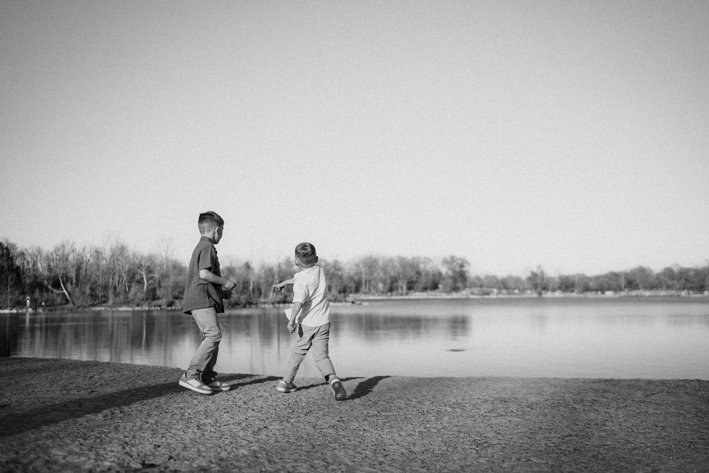 Two children playing with sticks along the shore of a peaceful lake on a calm day in black and white.