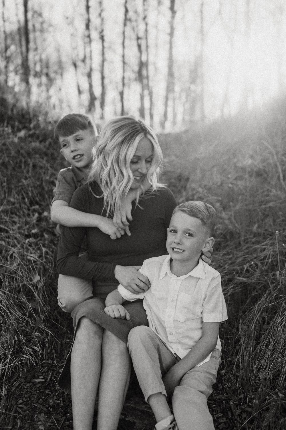 A black and white portrait of a parent and two children sitting together outdoors in tall grass with sunlight behind them.