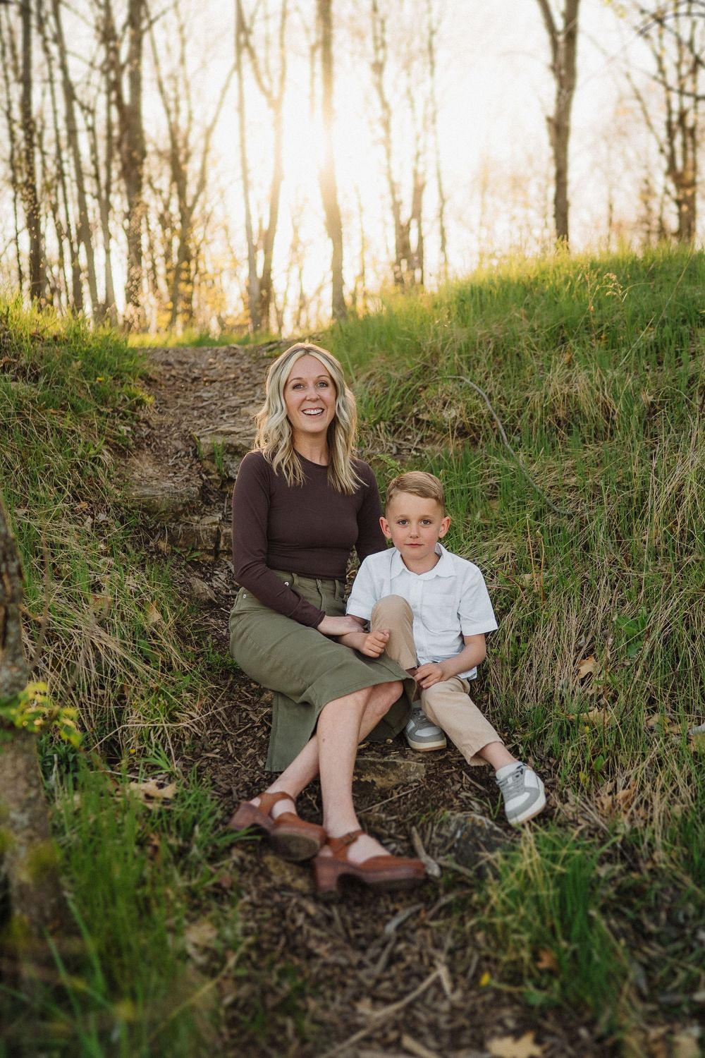 Parent and child sit together on a sunlit forest trail surrounded by tall trees and green grass.