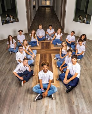 A group of young people wearing white shirts and blue jeans arranged in a cross formation on a wooden floor hallway.