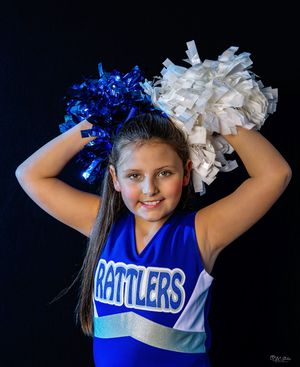 Cheerleader in blue uniform holding blue and white pom poms against black background.