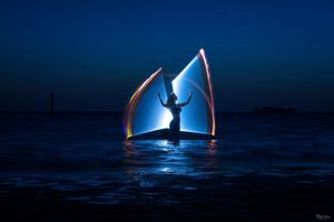Glowing sailboat illuminated by moonlight creates ethereal reflections on calm dark ocean waters at night.