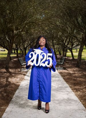 Graduate in blue cap and gown holds 2025 numbers while standing on pathway surrounded by trees.