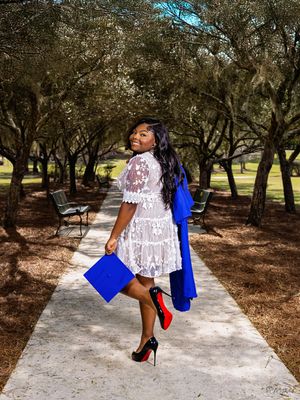 Graduate in white lace dress and blue graduation gown spins joyfully on a tree-lined path.