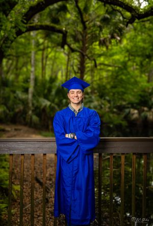 Graduate in blue cap and gown stands on wooden deck in forest setting.