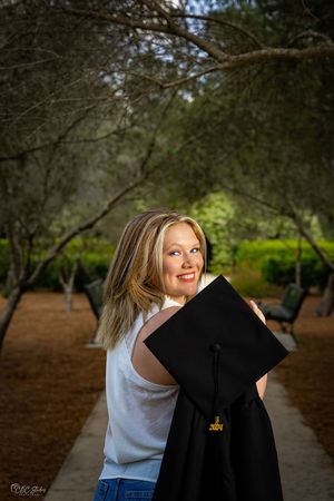 Casual graduate portrait holding black graduation cap in outdoor park setting.