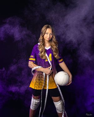 Volleyball player in purple and gold uniform holds ball and net against dark background.