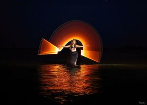 A silhouette dances on water against a glowing orange light circle created by steel wool spinning in a long exposure photo.