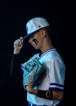 Baseball player in white uniform and blue cap poses dramatically against dark background with spotlight.