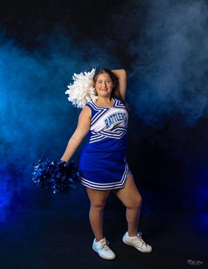 Cheerleader in blue and white uniform poses with pom poms against smoky blue backdrop.