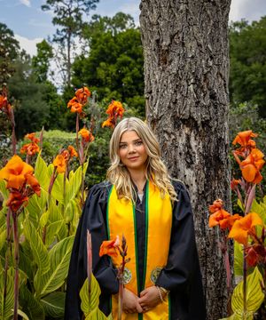 Graduate poses in cap and gown among orange flowers in garden setting.