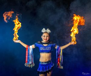 Cheerleader in blue uniform holds flaming pom poms against dark background.