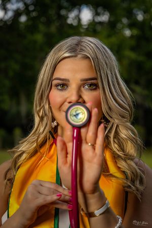 Graduate in yellow stole holds diploma while smiling against garden backdrop.