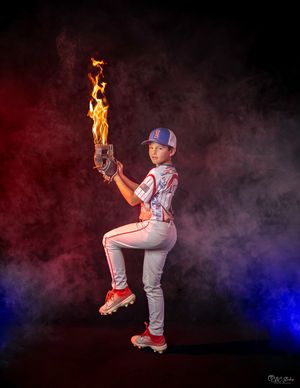 Baseball player in white uniform striking dramatic pose with flame effect in smoky studio setting.