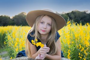 A person with a beige hat holds yellow flowers while posing in a field of wildflowers.