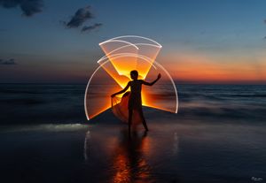 Silhouette performing light painting creating geometric patterns against ocean sunset.