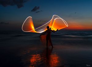 Person creating light trails with LED tools against beach sunset creating wing-like patterns.